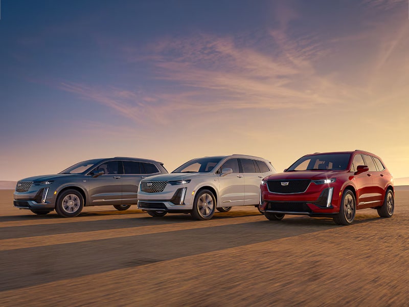 Three SUVs and a pickup truck are lined up. From left to right, a white SUV with a circular logo, a white SUV and a black pickup truck, both with GMC logos. All vehicles are shown from the front at a slight angle.